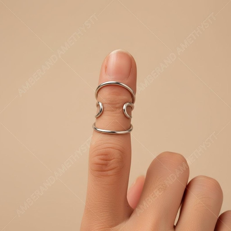 May include: A silver finger ring with a distinctive design. The ring features two bands linked by curved details, offering a modern, minimalist aesthetic. The ring is displayed on a finger against a plain backdrop.