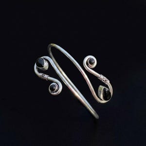 May include: A silver-toned open cuff bracelet featuring a bypass design with two spiraling ends. Each end is accented with a small, smooth, black oval stone, and the metal near the spirals displays a textured, decorative wire-wrapped detail.