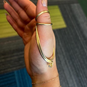 May include: A gold-colored ring set. The ring set features a thumb ring, a finger ring, and a curved design that connects the two rings. A delicate gold bracelet is also visible.