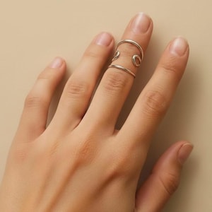 May include: A silver finger ring with an open design. The ring features two bands connected by curved and circular elements. The ring is worn on a finger against a neutral background.