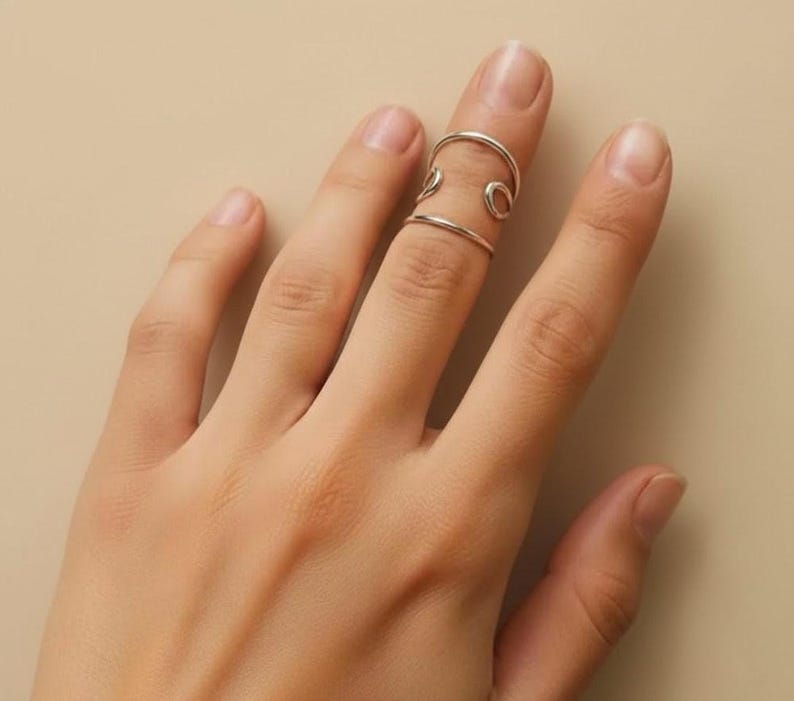 May include: A silver finger ring with an open design. The ring features two bands connected by curved and circular elements. The ring is worn on a finger against a neutral background.