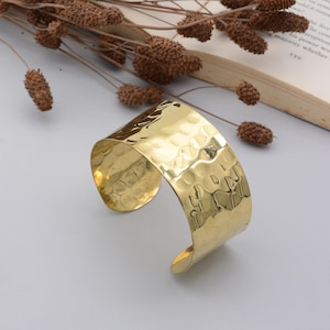 May include: A wide, hammered-gold cuff bracelet. The bracelet has a textured surface with a series of small, irregular indentations. The cuff is open at the bottom and has rounded edges. The background includes dried plants and a book.