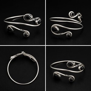 May include: A silver-toned open cuff bracelet featuring two curved ends, each accented with a smooth, teardrop-shaped black stone. The metal band displays intricate etched detailing near the stone settings, creating a balanced, symmetrical design.