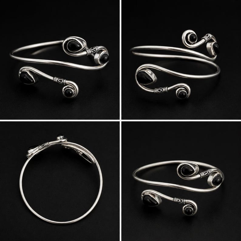 May include: A silver-toned open cuff bracelet featuring two curved ends, each accented with a smooth, teardrop-shaped black stone. The metal band displays intricate etched detailing near the stone settings, creating a balanced, symmetrical design.