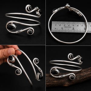 May include: A silver bypass arm cuff bracelet featuring four black teardrop-shaped stones set at the ends of spiraled metal terminals. A metal ruler placed alongside the bracelet indicates a diameter of approximately 3 inches across the band.