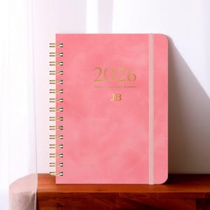 May include: A pink 2026 weekly and monthly planner with gold lettering and a spiral binding. The cover features the year 2026, the initials "JB", and a matching elastic band closure. The planner is displayed on a wooden surface.