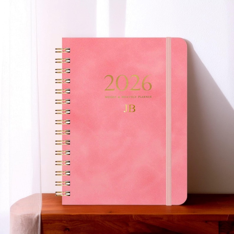 May include: A pink 2026 weekly and monthly planner with gold lettering and a spiral binding. The cover features the year 2026, the initials "JB", and a matching elastic band closure. The planner is displayed on a wooden surface.