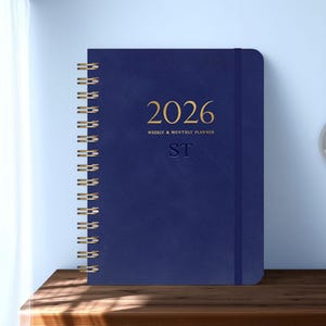 May include: A navy blue 2026 weekly and monthly planner with gold spiral binding. The cover features the year 2026 in gold lettering, along with the text "WEEKLY & MONTHLY PLANNER" and the initials "ST". The planner is closed with an elastic band.