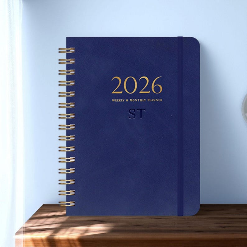 May include: A navy blue 2026 weekly and monthly planner with gold spiral binding. The cover features the year 2026 in gold lettering, along with the text "WEEKLY & MONTHLY PLANNER" and the initials "ST". The planner is closed with an elastic band.