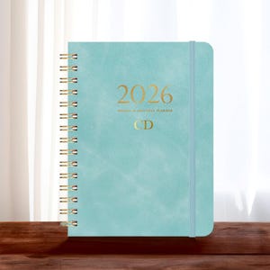 May include: A teal 2026 weekly and monthly planner with gold spiral binding and elastic closure. The cover features the year 2026 and initials "CD" in gold lettering. The planner is on a wooden surface.