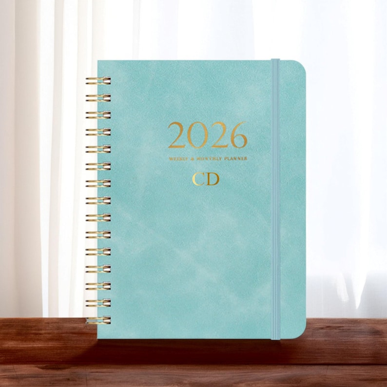 May include: A teal 2026 weekly and monthly planner with gold spiral binding and elastic closure. The cover features the year 2026 and initials "CD" in gold lettering. The planner is on a wooden surface.