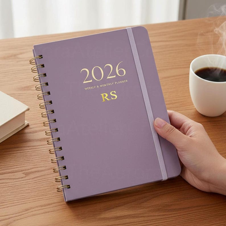 May include: A lavender 2026 weekly and monthly planner with gold lettering and a spiral binding. The planner has an elastic band closure and the initials "RS" are printed on the cover. A white mug of coffee sits nearby.