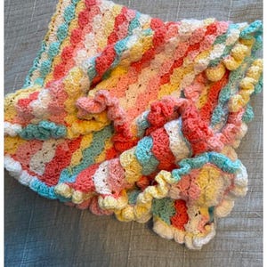 May include: A colorful, handmade crochet blanket with a scalloped edge. The blanket features stripes of pink, yellow, blue, and white. The edges are trimmed with a white, ruffled border. The blanket is folded and draped on a gray surface.