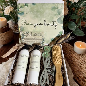May include: A gift box with hair care products, including two white bottles of shampoo and conditioner, a wooden hairbrush, and hair clips. The box also contains small candles and a card with the text "Own your beauty".