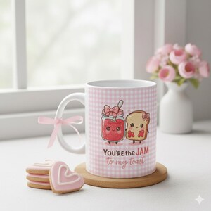 Puede incluir: Una taza de cerámica blanca con un estampado de cuadros rosas y una ilustración de dibujos animados de mermelada y tostadas. La taza tiene el texto "You're the JAM to my toast". Una cinta rosa está atada al asa. Galletas en forma de corazón en primer plano.