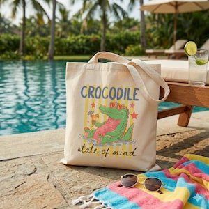 Puede incluir: Un bolso tote de lona beige con un gráfico colorido de un cocodrilo de dibujos animados con gafas de sol y un cóctel. El texto "CROCODILE state of mind" está impreso encima y debajo de la ilustración. El bolso está junto a una piscina.