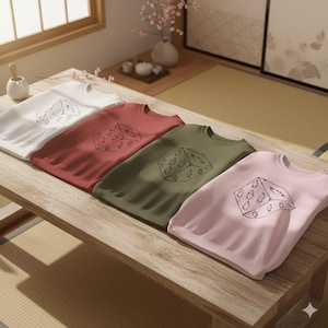 May include: Four crewneck sweatshirts in white, red, olive green, and pink, each featuring a black line drawing of a die. The sweatshirts are arranged on a wooden table, with a minimalist aesthetic. The image is well-lit, showcasing the colors and design.