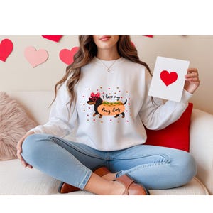 Puede incluir: Sudadera blanca con un diseño de perro salchicha con gafas de corazón y el texto "I love my long dog". El perro está diseñado para parecerse a un perrito caliente. Pequeños corazones rojos están dispersos alrededor del diseño. Una persona sostiene una tarjeta con un corazón rojo.
