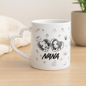Puede incluir: Taza de cerámica blanca con asa en forma de corazón. La taza presenta una ilustración en blanco y negro de estilo manga de dos mujeres dentro de corazones, con la palabra "NANA" debajo. Pequeños dibujos de fresas y otros objetos están dispersos.