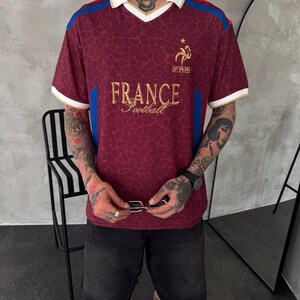 May include: A burgundy short-sleeve polo shirt with a white collar and trim. The shirt features a repeating giraffe-like pattern, blue side panels, and gold embroidery on the chest that reads FRANCE Football with a rooster emblem above it.