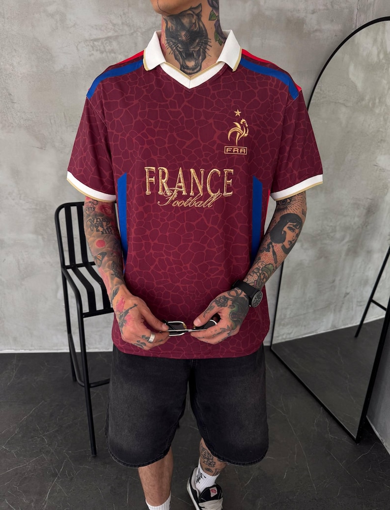 May include: A burgundy short-sleeve polo shirt with a white collar and trim. The shirt features a repeating giraffe-like pattern, blue side panels, and gold embroidery on the chest that reads FRANCE Football with a rooster emblem above it.