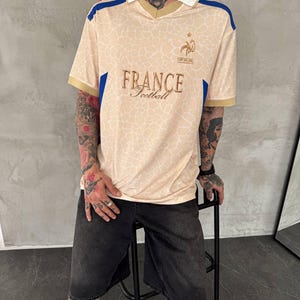 May include: A beige short-sleeved jersey with a white collar and a subtle giraffe-like pattern. The shirt features blue and red shoulder accents, a gold embroidered rooster emblem, and the text FRANCE Football printed in gold on the chest.