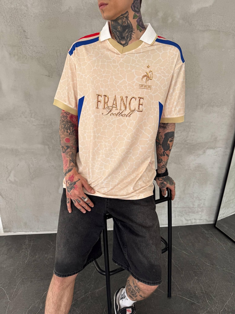 May include: A beige short-sleeved jersey with a white collar and a subtle giraffe-like pattern. The shirt features blue and red shoulder accents, a gold embroidered rooster emblem, and the text FRANCE Football printed in gold on the chest.