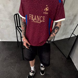 May include: A maroon V-neck jersey with a white collar and gold trim. The shirt features a gold rooster emblem above the text "FRANCE football" and "FRA". It has a dark, web-like pattern across the fabric, with blue and red stripes on the shoulders.
