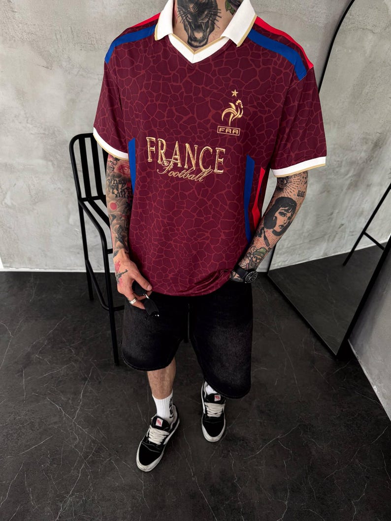 May include: A maroon V-neck jersey with a white collar and gold trim. The shirt features a gold rooster emblem above the text "FRANCE football" and "FRA". It has a dark, web-like pattern across the fabric, with blue and red stripes on the shoulders.