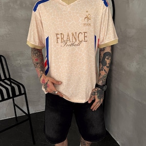 May include: A beige short-sleeved polo shirt with a white collar and gold trim. The shirt features a giraffe-like pattern, blue and red side stripes, and the text "FRANCE Football" in gold embroidery on the chest, along with a small logo above it.