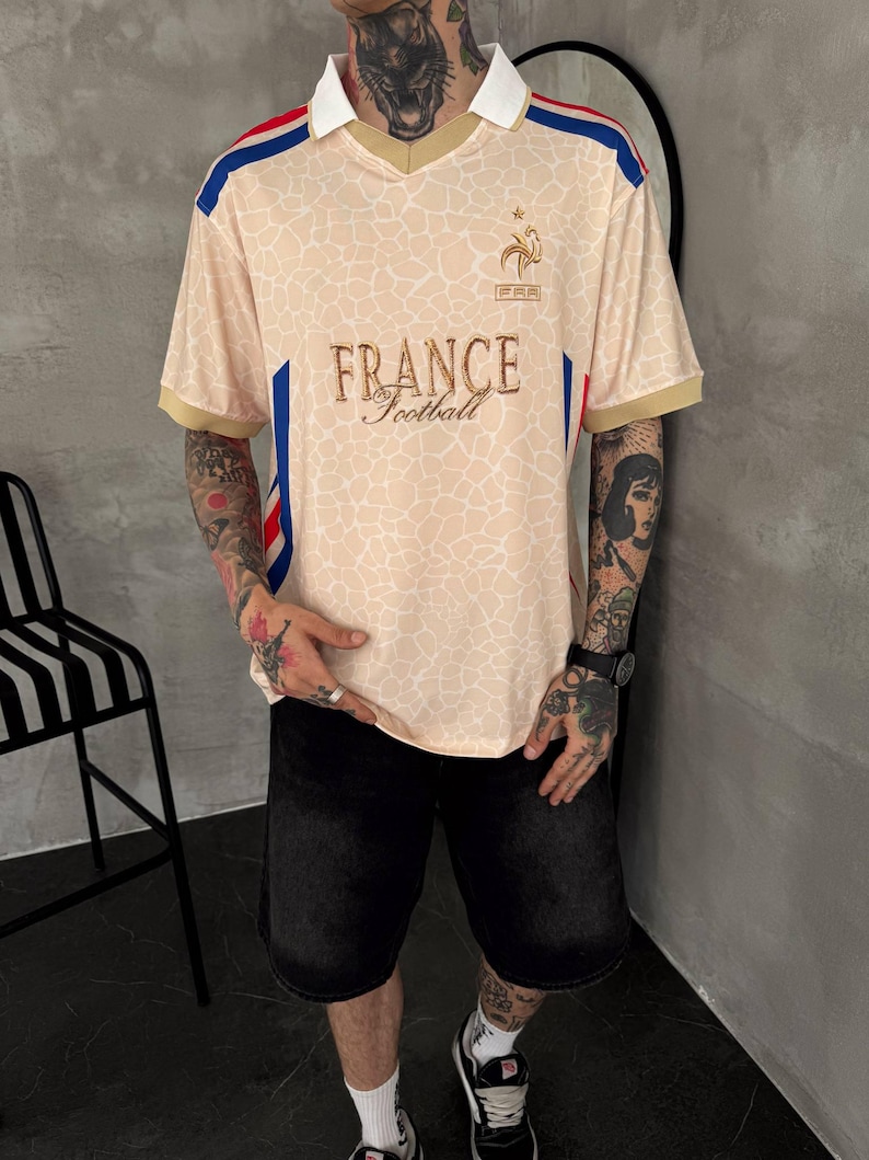 May include: A beige short-sleeved polo shirt with a white collar and gold trim. The shirt features a giraffe-like pattern, blue and red side stripes, and the text "FRANCE Football" in gold embroidery on the chest, along with a small logo above it.