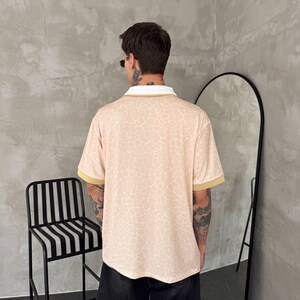May include: A short-sleeved polo shirt featuring a light peach base with a white crackle or giraffe-like pattern, a solid white collar, and solid tan sleeve cuffs, paired with black shorts.