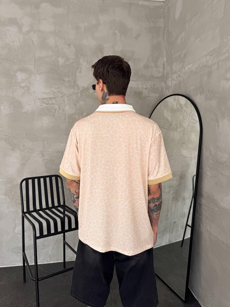 May include: A short-sleeved polo shirt featuring a light peach base with a white crackle or giraffe-like pattern, a solid white collar, and solid tan sleeve cuffs, paired with black shorts.