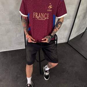May include: A maroon short-sleeved jersey with a white collar, featuring a gold France Football logo and text on the chest. The fabric has a subtle tonal giraffe-like pattern, with blue and red accents on the shoulders and white trim on the sleeves.