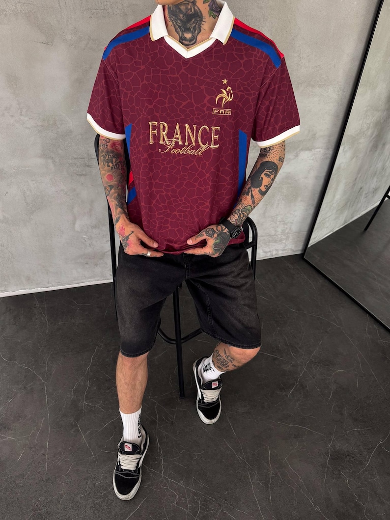 May include: A maroon short-sleeved jersey with a white collar, featuring a gold France Football logo and text on the chest. The fabric has a subtle tonal giraffe-like pattern, with blue and red accents on the shoulders and white trim on the sleeves.