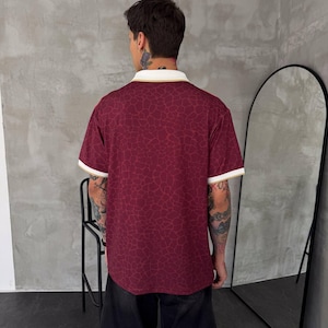 May include: A short-sleeved polo shirt in a deep burgundy color featuring a subtle, darker red giraffe-like pattern throughout. The shirt has a white collar and sleeve cuffs, both accented with a thin gold stripe along the edges.