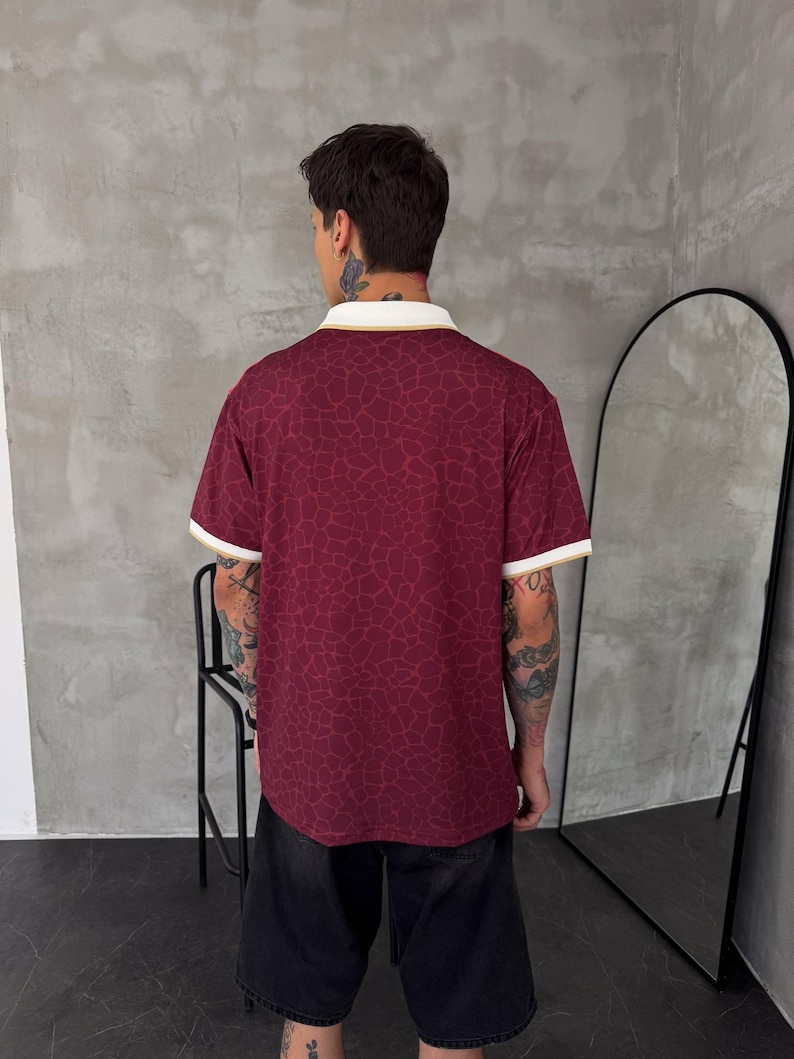 May include: A short-sleeved polo shirt in a deep burgundy color featuring a subtle, darker red giraffe-like pattern throughout. The shirt has a white collar and sleeve cuffs, both accented with a thin gold stripe along the edges.