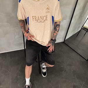 May include: A beige short-sleeved jersey with a white collar, featuring a subtle giraffe-like pattern, blue and red shoulder stripes, and the text "FRANCE Football" with a gold rooster emblem on the chest, paired with black denim shorts and sneakers.