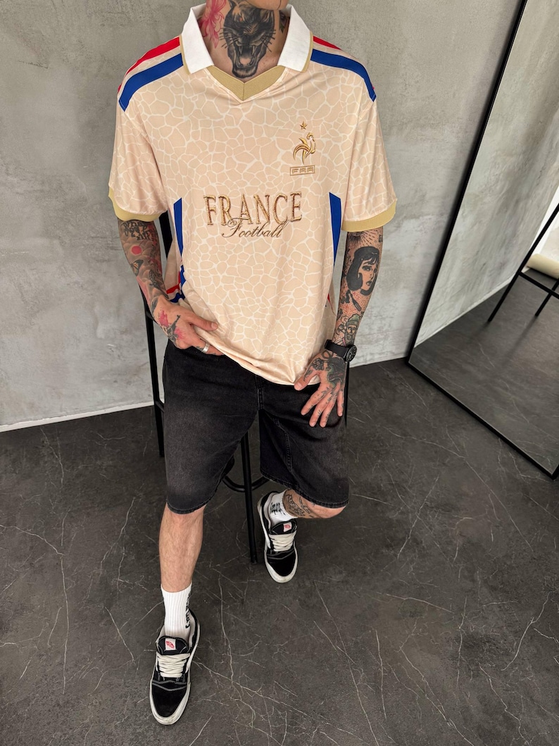May include: A beige short-sleeved jersey with a white collar, featuring a subtle giraffe-like pattern, blue and red shoulder stripes, and the text "FRANCE Football" with a gold rooster emblem on the chest, paired with black denim shorts and sneakers.