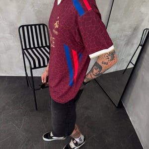 May include: A maroon short-sleeved jersey with a subtle giraffe-like pattern, featuring a white collar, white sleeve cuffs, and vertical blue and red stripes on the shoulders and sides. A gold emblem and the text FFF are visible on the chest.