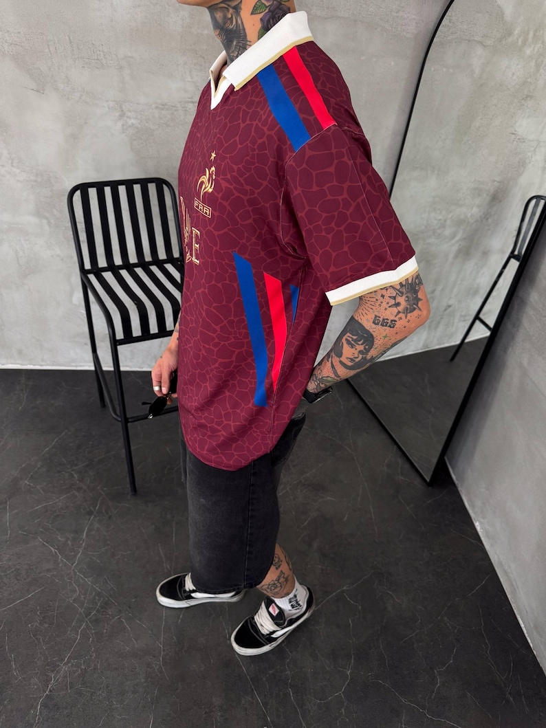 May include: A maroon short-sleeved jersey with a subtle giraffe-like pattern, featuring a white collar, white sleeve cuffs, and vertical blue and red stripes on the shoulders and sides. A gold emblem and the text FFF are visible on the chest.