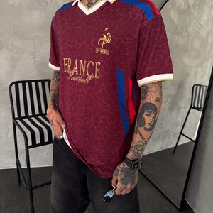 May include: A maroon short-sleeved jersey with a white collar and gold trim. The shirt features a gold rooster emblem above the text "FRA" and "FRANCE Football" in gold lettering. It has a giraffe-like pattern and blue and red side stripes.