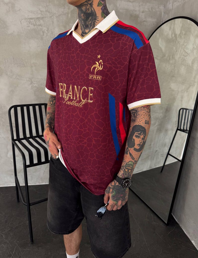 May include: A maroon short-sleeved jersey with a white collar and gold trim. The shirt features a gold rooster emblem above the text "FRA" and "FRANCE Football" in gold lettering. It has a giraffe-like pattern and blue and red side stripes.