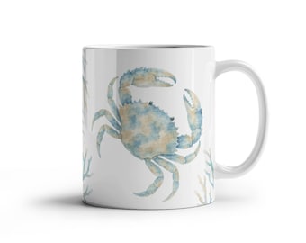 Puede incluir: Una taza de cerámica blanca con una ilustración en acuarela de un cangrejo azul y bronceado centrado en el frente, rodeado de representaciones más pequeñas y tenues de ramas de coral azul y bronceado en los lados del recipiente.