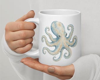 Puede incluir: Una taza de cerámica blanca con una ilustración estilo acuarela de un pulpo en tonos moteados azul y beige. El pulpo se muestra con los tentáculos curvados hacia afuera, centrado en el lateral de la taza cilíndrica de superficie lisa.