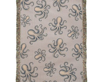 Puede incluir: Una manta tejida con un patrón repetitivo de pulpos azules y beige sobre un fondo gris claro. La manta rectangular tiene una textura de tela tejida y está terminada con flecos decorativos a lo largo de los cuatro bordes.