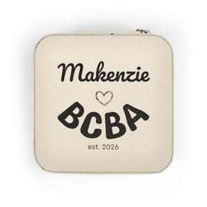 Könnte beinhalten: Ein quadratisches, cremefarbenes Schmuckkästchen mit Reißverschluss. Das Etui trägt den Namen "Makenzie" in schwarzer Schrift über einem Herz und die Buchstaben "BCBA" in einer fetten, geschwungenen Schrift. Darunter ist der Text "est. 2026" in Schwarz gedruckt.