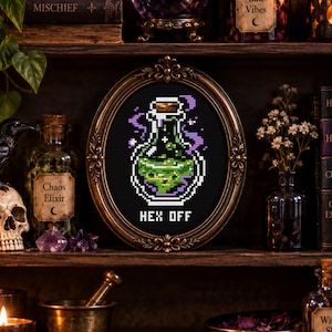 May include: A cross-stitch embroidery in an oval bronze-toned frame featuring a pixelated glass bottle filled with a glowing green liquid. Purple smoke swirls around the bottle, and the text "HEX OFF" is stitched in white below the bottle.