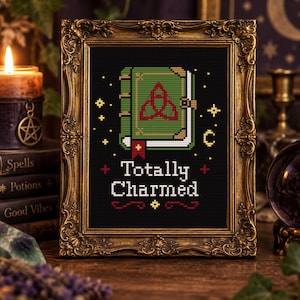 May include: A cross-stitch pattern in a gold ornate frame featuring a green book with a red triquetra symbol on the cover. The text "Totally Charmed" is stitched in white below the book, surrounded by small yellow stars and red decorative accents.