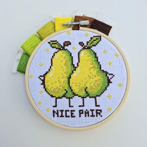 May include: A cross-stitch embroidery in a wooden hoop depicts two yellow and green pears with the text "NICE PAIR". Embroidery threads in green, yellow, and brown are visible above the hoop.